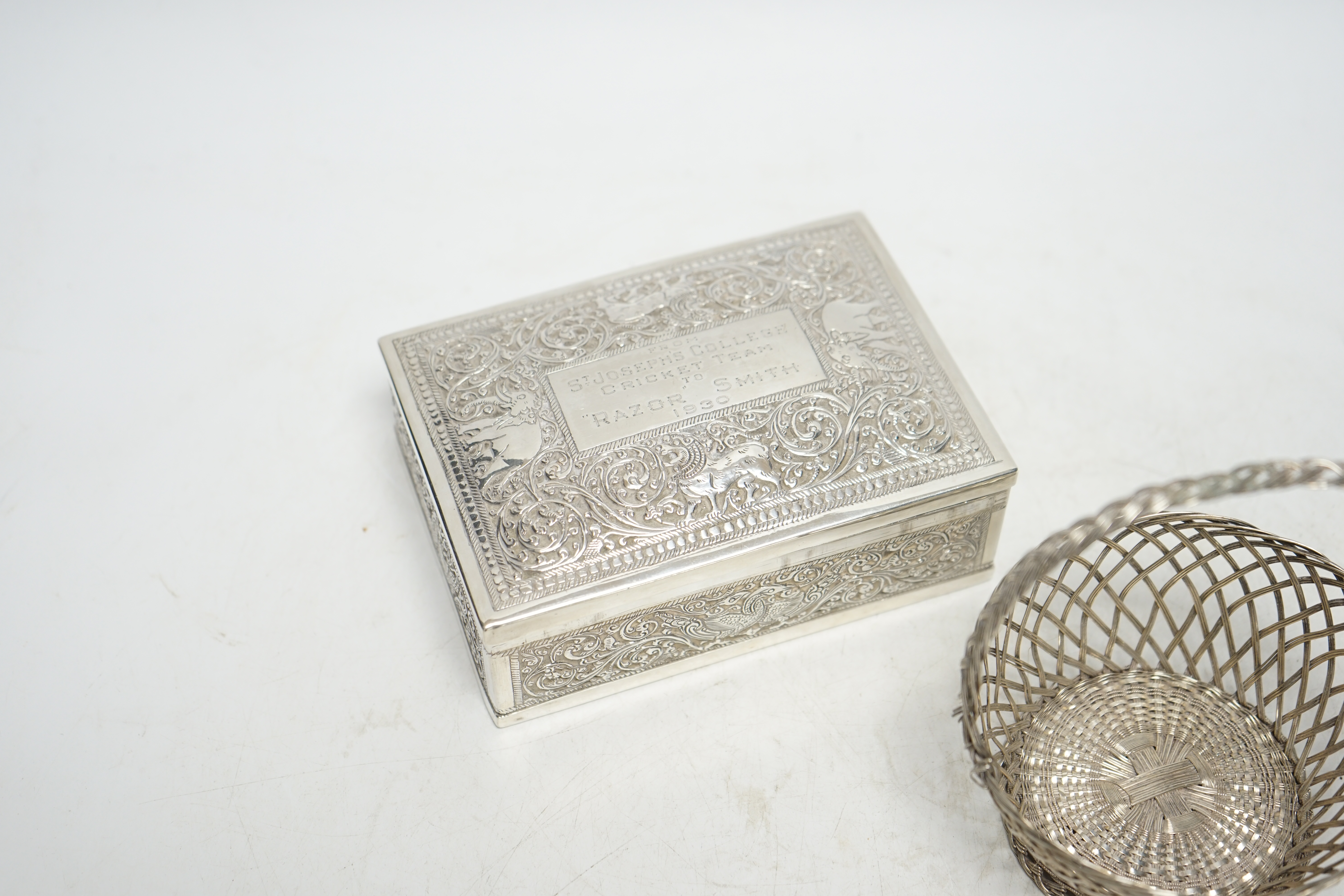 An Indian white metal mounted rectangular cigarette box, with engraved inscription, 'From St. Joseph's College Cricket Team, to 'Razor' Smith, 1930', 12.9cm, together with a white metal open work basket. Condition - fair